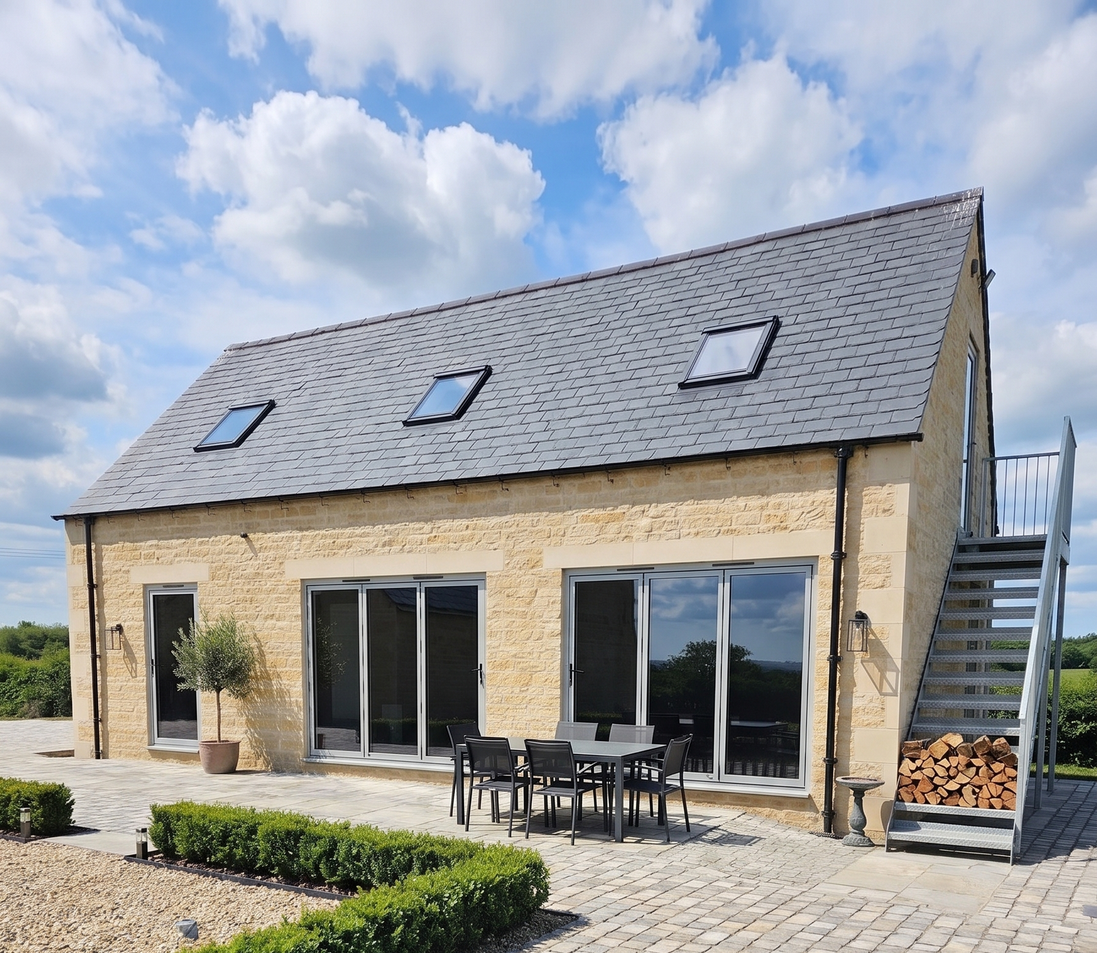 A modern single-storyhome gym in stone with a gray slate roof, large glass sliding doors, a paved patio, outdoor dining furniture, a small staircase, and landscaped hedges under a blue sky with clouds.  